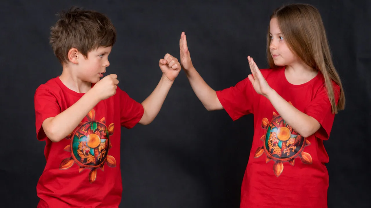 Zwei Kinder in roten WingTsun-Shirts stehen sich gegenüber; das Mädchen hält die offene Hand als klares Stopp vor sich.